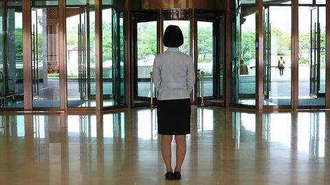 Frau steht in einem Foyer und blickt durch den gläsernen Eingangsbereich nach draußen.