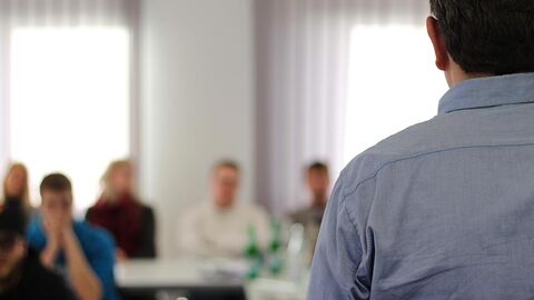 Unterricht (Symbolbild) Lehrer steht vor Auszubildenden, die nur unscharf zu erkennen sind.