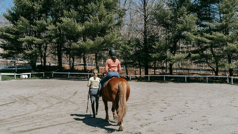 Reitlehrerin führt eine Schülerin auf einem Pferd über einen Reitplatz.
